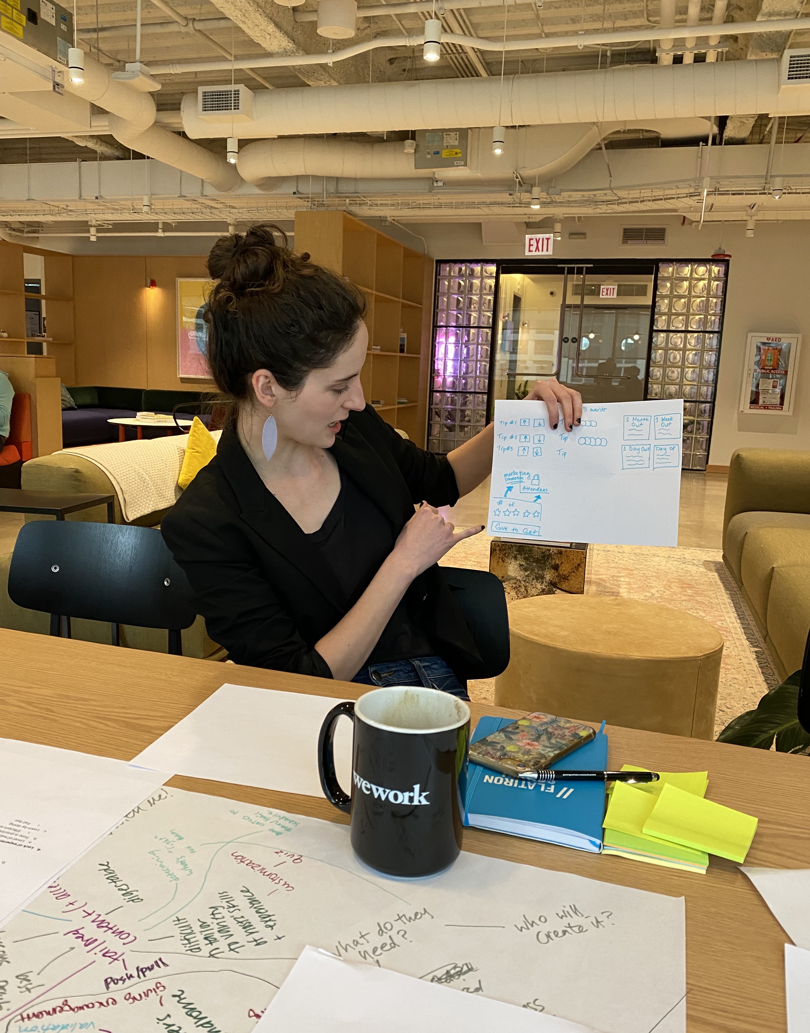 Image of the author holding up a sketch while talking through it. There's a table in the foreground, littered with post-it notes, papers, markers, and coffee cups.